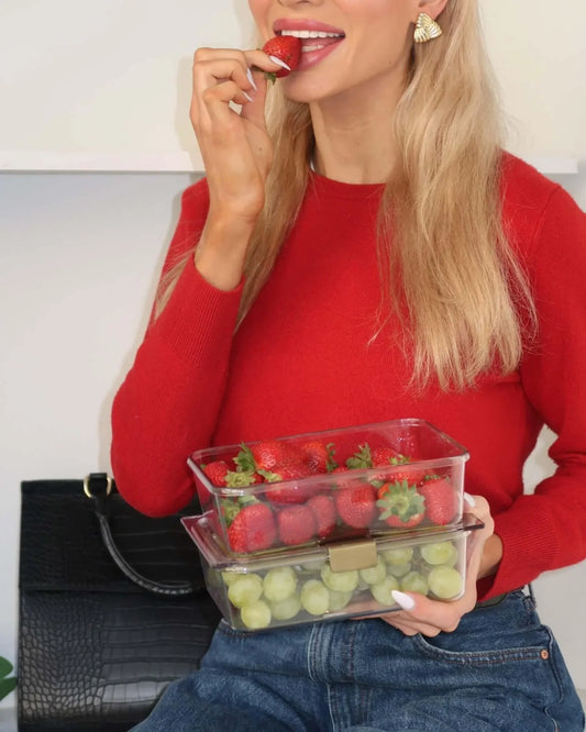 Woman in red sweater eating strawberry, holding clear lunch containers with grapes and strawberries, black vegan leather tote beside her