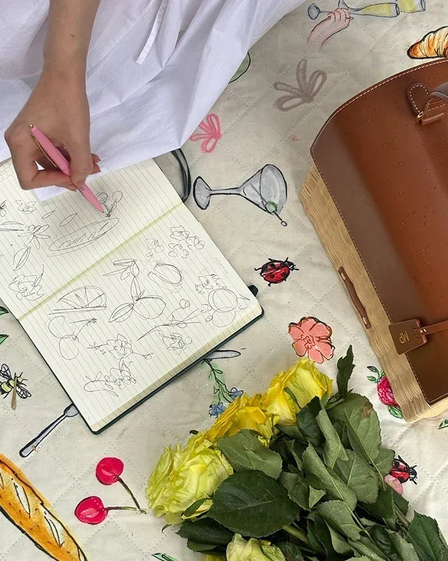 Sketching floral designs beside a chic brown vegan leather tote, yellow roses, and a picnic-themed blanket.