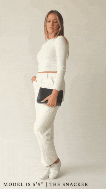 A full-body animation shows a blonde woman wearing a white long-sleeve top and white trousers modeling the small, rectangular pouch. She holds the black pouch by her side, then the animation shows her opening the zipper to reveal the interior.