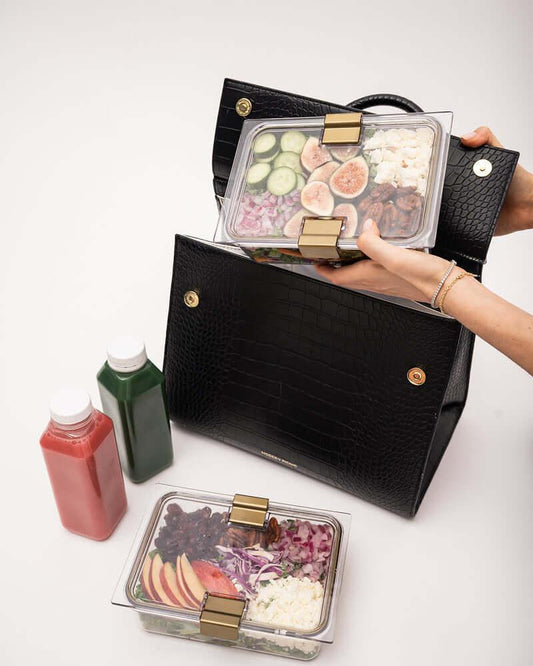 Black textured lunch tote with gold clasps holding healthy meal prep containers and juice bottles