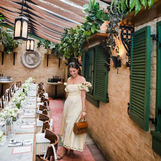 Woman in a cream dress holds a chic lunch tote in a stylish, plant-filled restaurant setting
