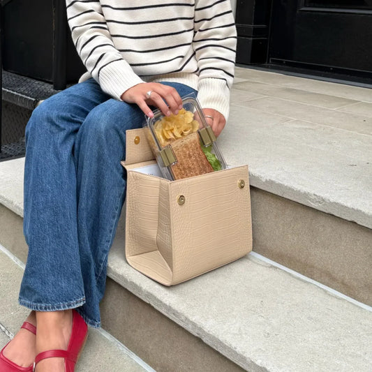 Woman sitting on steps placing lunch container with sandwich and chips into beige vegan leather lunch bag