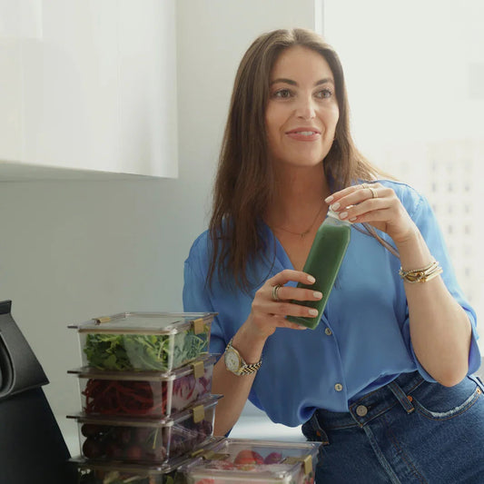 Woman in blue blouse holding green smoothie next to stacked salad lunch containers in modern kitchen