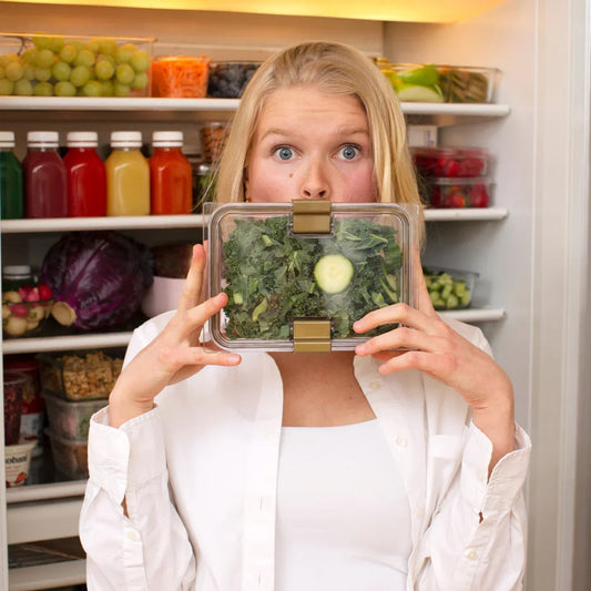 Woman holding salad container in front of face, open refrigerator with healthy meal prep.
