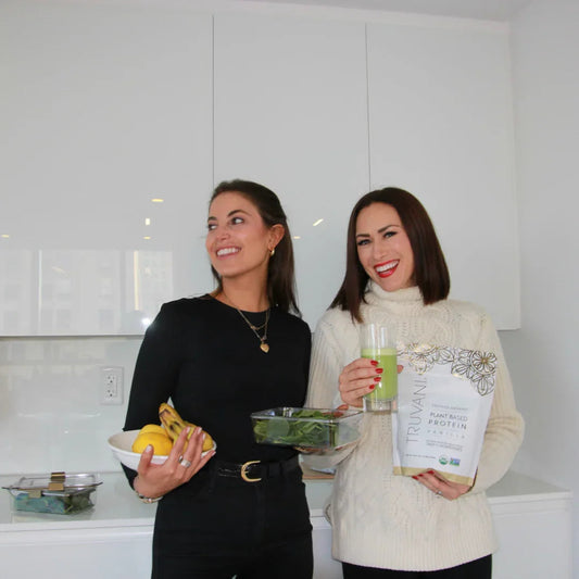 Two women in a modern kitchen holding healthy food, fresh produce, and a plant protein shake