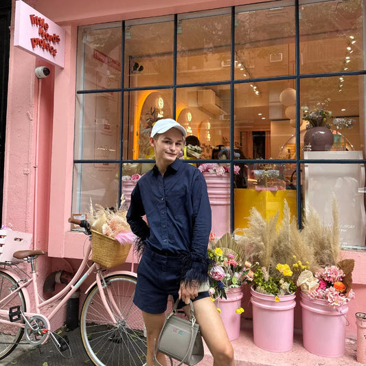 Person in navy outfit with white cap posing by pink bicycle and flower buckets outside shop.
