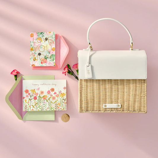 Chic insulated wicker lunch bag with white flap, floral Mother’s Day cards, and pink flowers on pink background.