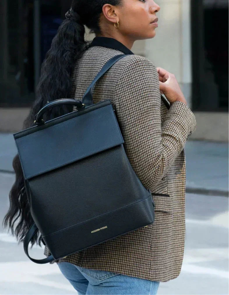 Black vegan leather backpack with top handle, worn by woman in houndstooth blazer for work or travel