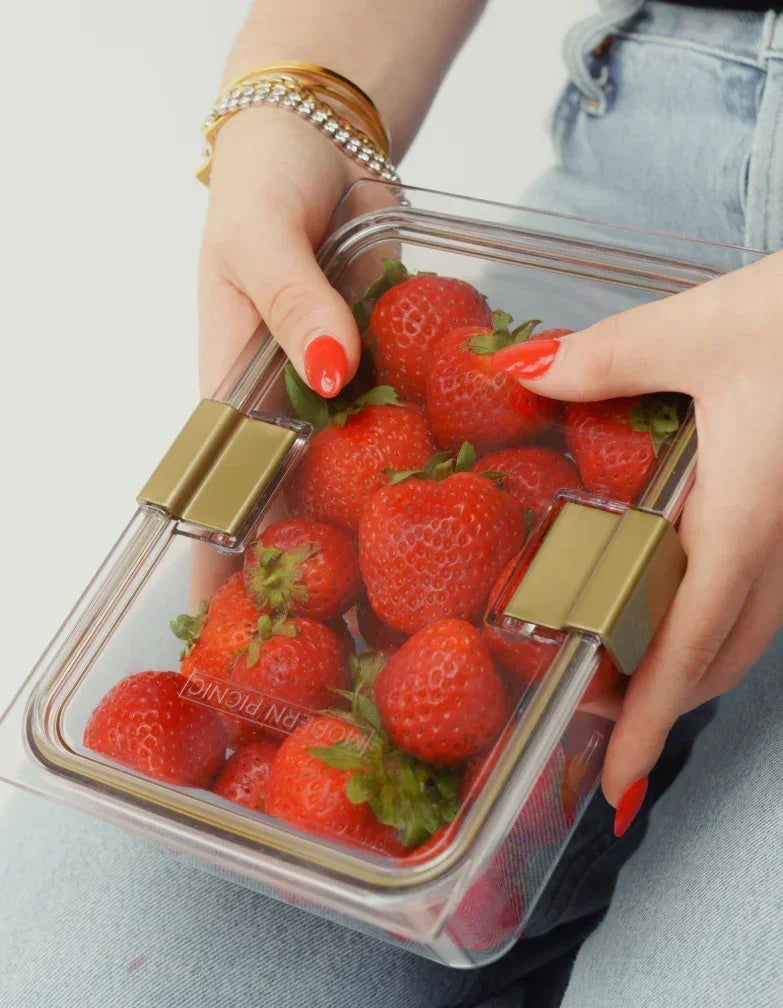 Clear lunch container with fresh strawberries, gold clasps, held by hands with red nails