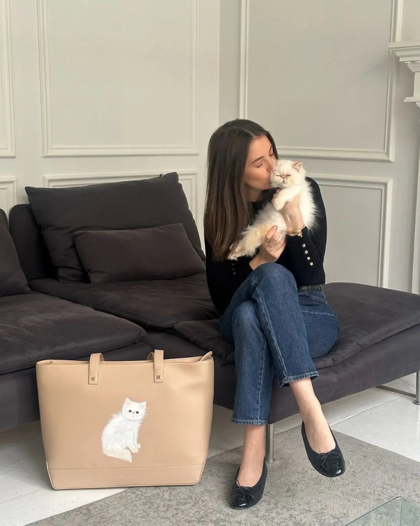 Woman sitting on sofa kissing white cat, beige vegan leather tote bag with cat design nearby