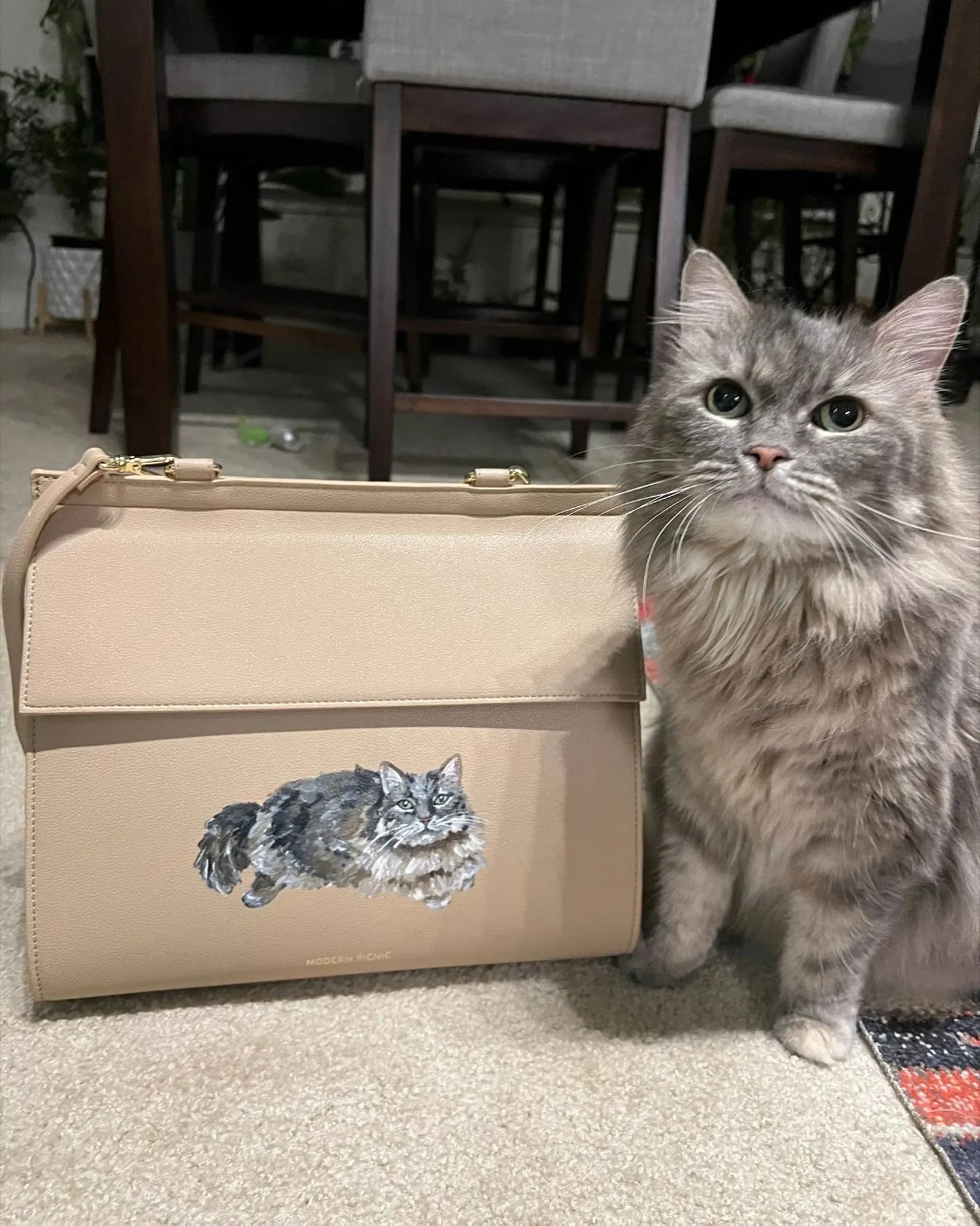 Gray long-haired cat beside tan vegan leather lunch bag with cat illustration