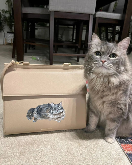 Gray fluffy cat next to beige vegan leather lunch bag with cat illustration, indoor setting