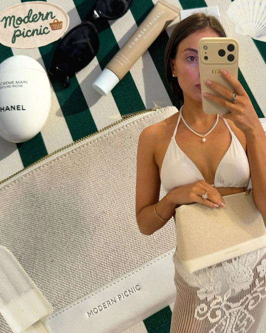 Stylish woman holding a chic beige clutch on a green-and-white striped picnic blanket with beauty essentials.