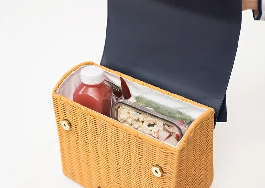 Open tan wicker insulated lunch bag containing a juice bottle and healthy meal prep containers.