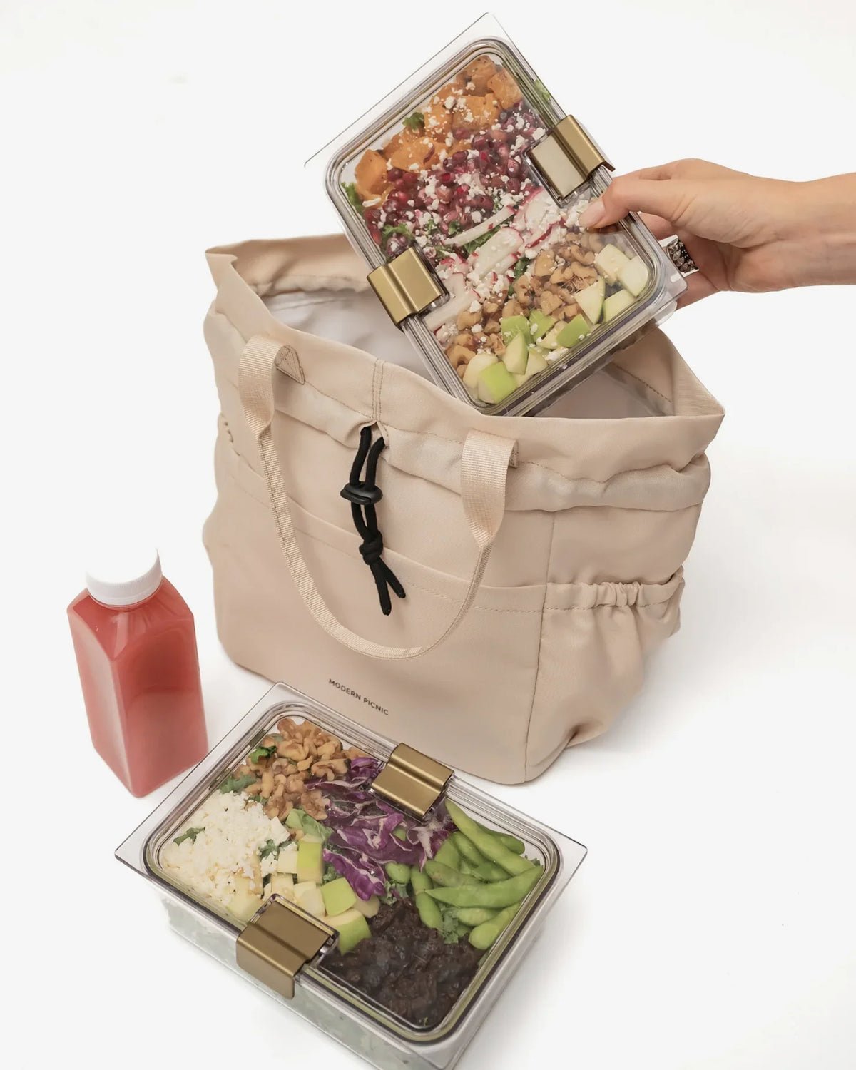 A person placing a clear glass meal prep container with a colorful salad into a beige insulated lunch tote bag.