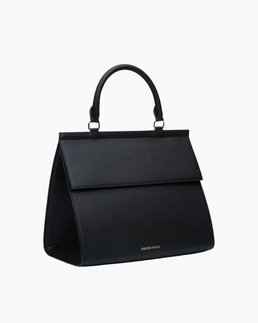 An angled studio shot of a black top-handle purse. The bag is structured, made of vegan leather, and stands upright, showing the depth of the rectangular body. The curved top handle and the gold MODERN PICNIC logo are visible on the front base.