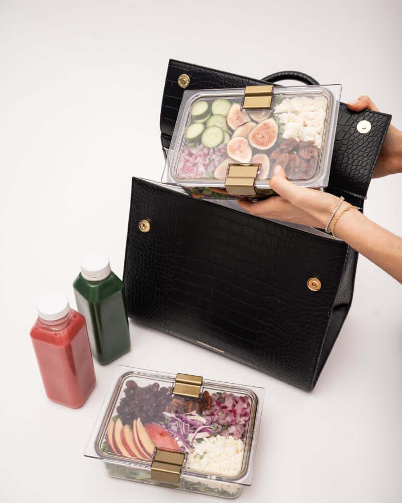 An overhead view of a woman packing a black crocodile-embossed handbag with a white insulated interior. One hand places a clear container holding a salad with figs, cucumber, and feta into the bag, while another container with a salad of apples, cabbage, and dried fruit rests below. Two juice bottles are also visible.