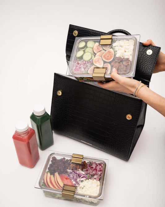 An overhead view of a woman packing a black crocodile-embossed handbag with a white insulated interior. One hand places a clear container holding a salad with figs, cucumber, and feta into the bag, while another container with a salad of apples, cabbage, and dried fruit rests below. Two juice bottles are also visible.