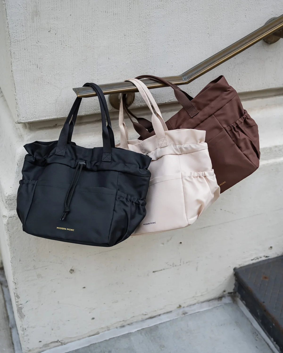 Three Modern Picnic insulated lunch totes in black, cream, and brown hanging on a handrail.