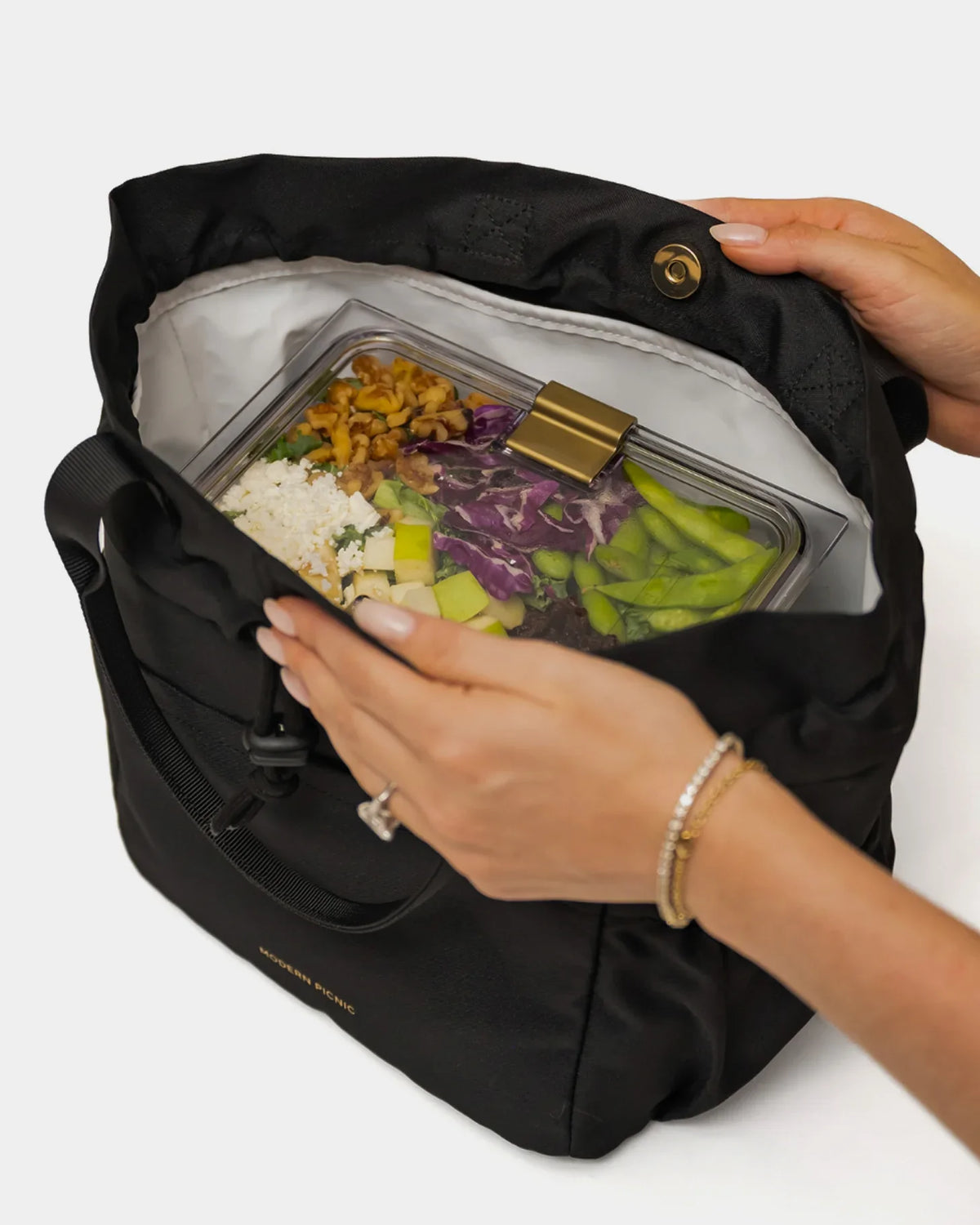 Hands opening a black insulated lunch tote containing a glass meal prep container with salad.
