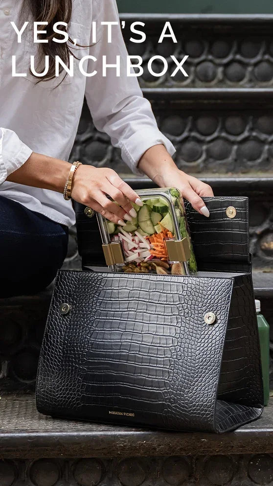 Woman placing a glass meal prep container with fresh vegetables into a black vegan leather lunch bag.