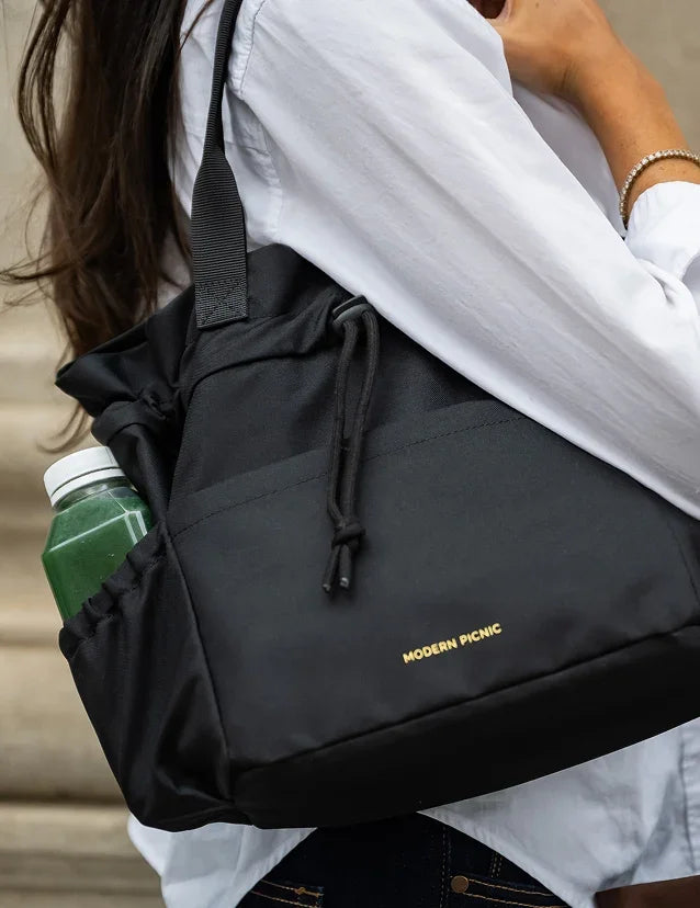 A person wearing a black Modern Picnic insulated tote bag with a green drink inside.