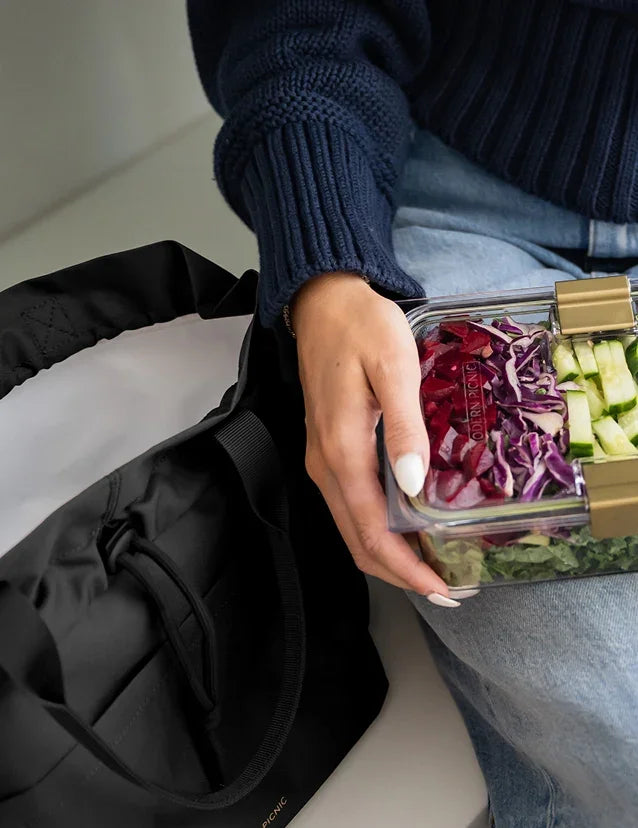 Person holding a clear glass meal prep container filled with fresh salad next to a black tote.