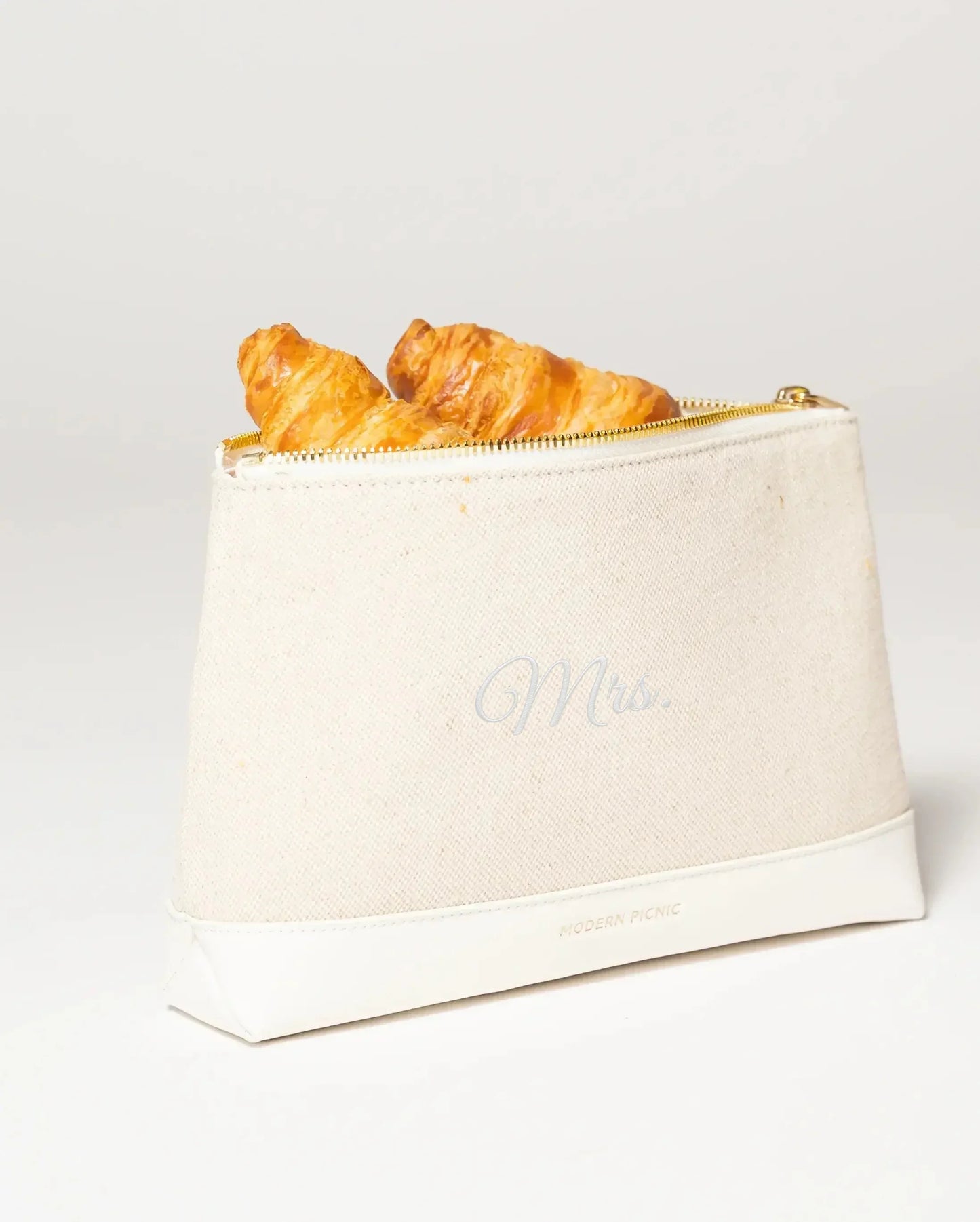 Cream canvas lunch bag with 'Mrs.' script, gold zipper, holding croissants, vegan leather base