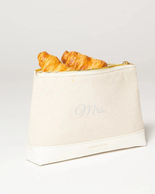 Cream canvas lunch bag with 'Mrs.' script, gold zipper, holding croissants, vegan leather base