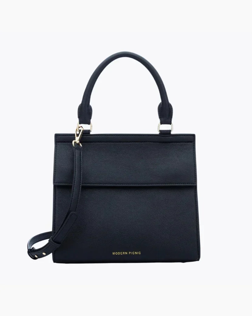 A front-facing studio shot of a structured black top-handle handbag. The bag is made of pebbled vegan leather and features a clean flap closure, a detachable matching shoulder strap, and gold hardware. The logo, "MODERN PICNIC," is subtly visible in gold near the bottom.
