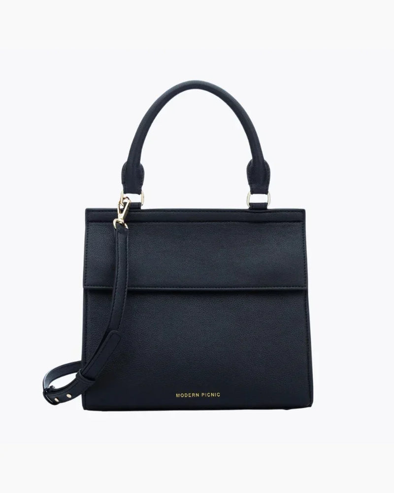 A front-facing studio shot of a structured black top-handle handbag. The bag is made of pebbled vegan leather and features a clean flap closure, a detachable matching shoulder strap, and gold hardware. The logo, "MODERN PICNIC," is subtly visible in gold near the bottom.