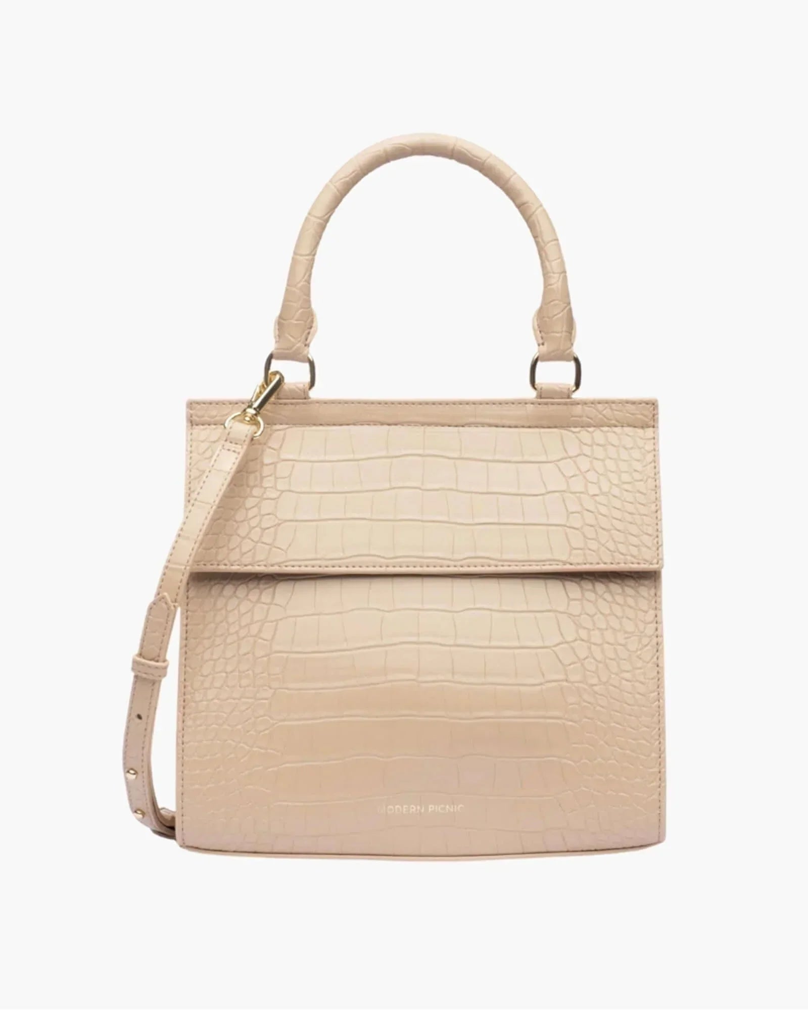 A front-facing studio shot of a structured, light beige top-handle handbag. The bag is covered in a crocodile-embossed texture and features a clean flap closure, a detachable shoulder strap, and gold hardware. The logo "MODERN PICNIC" is subtly visible in gold near the bottom.