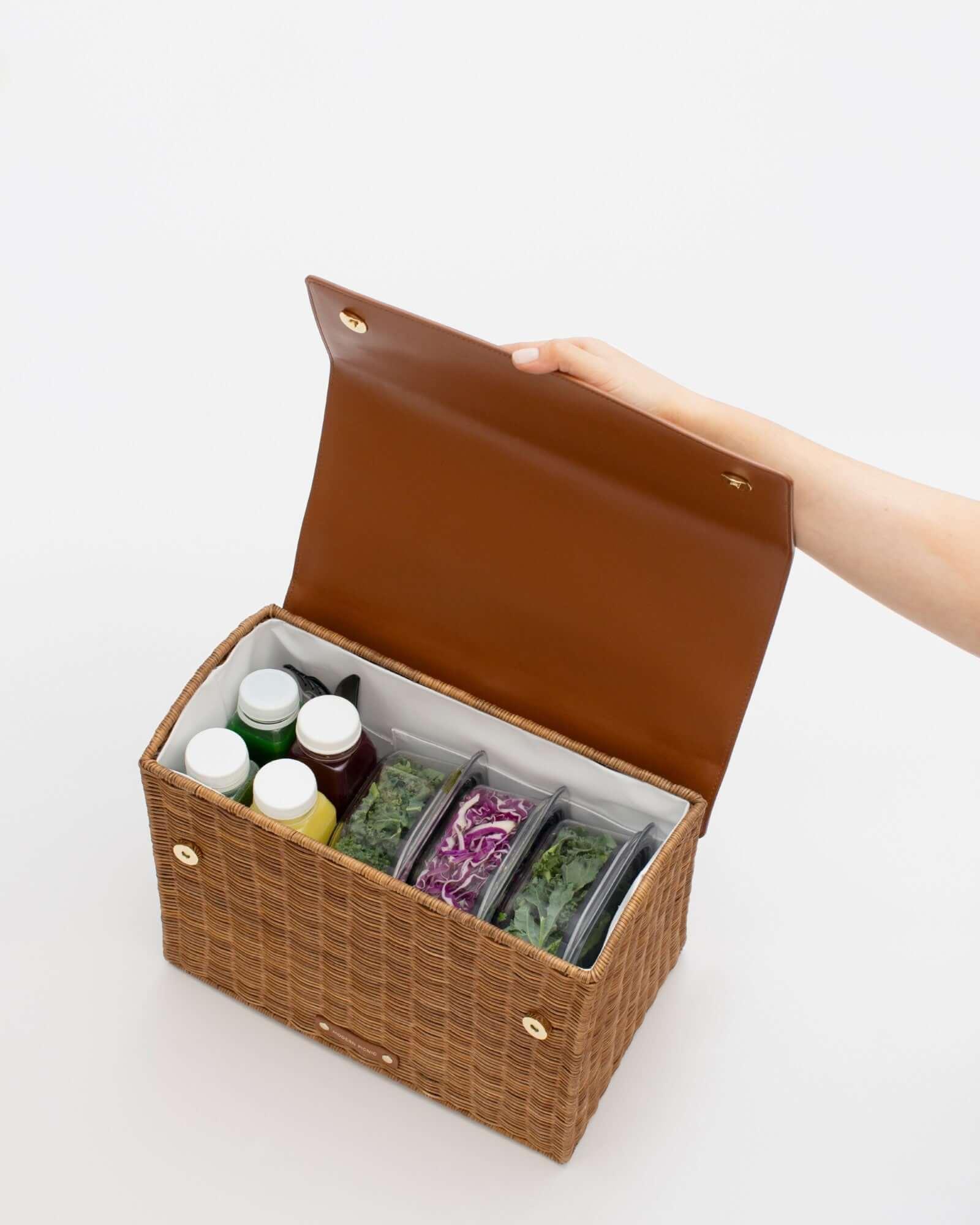 An overhead shot shows the picnic-style handbag with its brown vegan leather flap held fully open by a hand. The body of the bag is made of brown woven wicker and features a white insulated interior. The bag is packed with four juice bottles and three clear food containers holding salad ingredients like kale and purple cabbage.