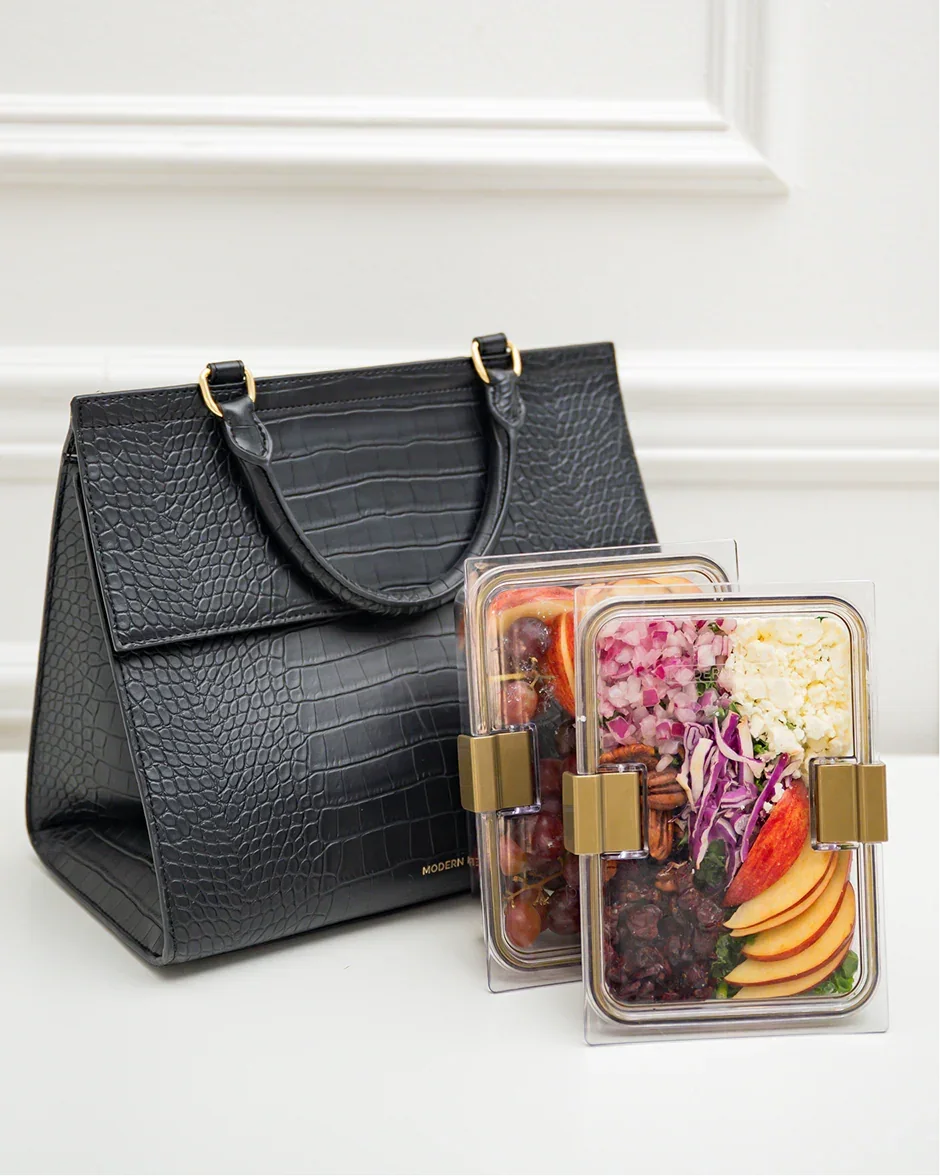 Black textured lunch tote with gold hardware beside clear bento boxes filled with colorful healthy meals