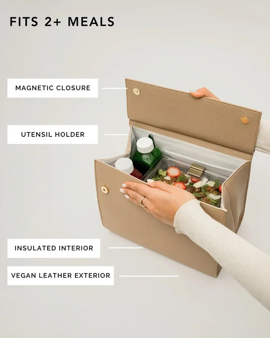 An overhead shot shows a cream-colored top-handle handbag opened to display its function as a lunch bag. Inside the main compartment, which has a white insulated lining, are two juice bottles and a clear container holding a salad with strawberries. Feature labels point to the magnetic closure, utensil holder (hidden under the flap), insulated interior, and vegan leather exterior. A hand with a gold ring is reaching into the bag. The text overlay reads, "FITS 2+ MEALS."