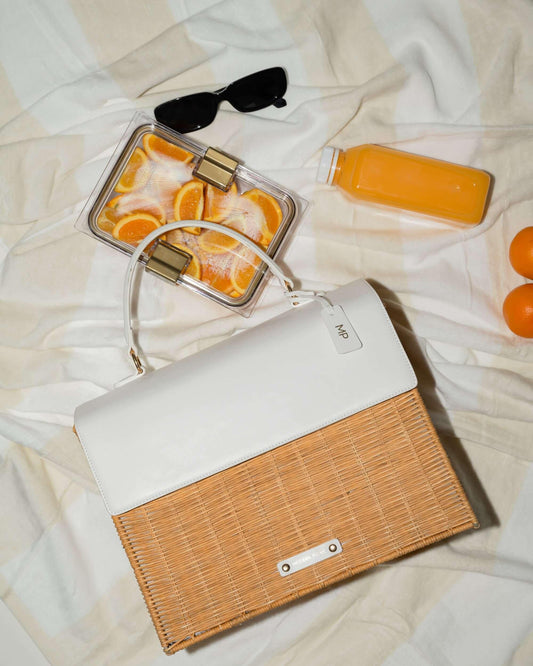 An overhead flat lay shot shows a picnic scene on a neutral-striped blanket. The main item is a structured handbag with a brown woven wicker body and a white vegan leather top flap. Around the bag are picnic accessories, including black sunglasses, a bottle of orange juice, a clear container of sliced oranges, and two whole oranges.