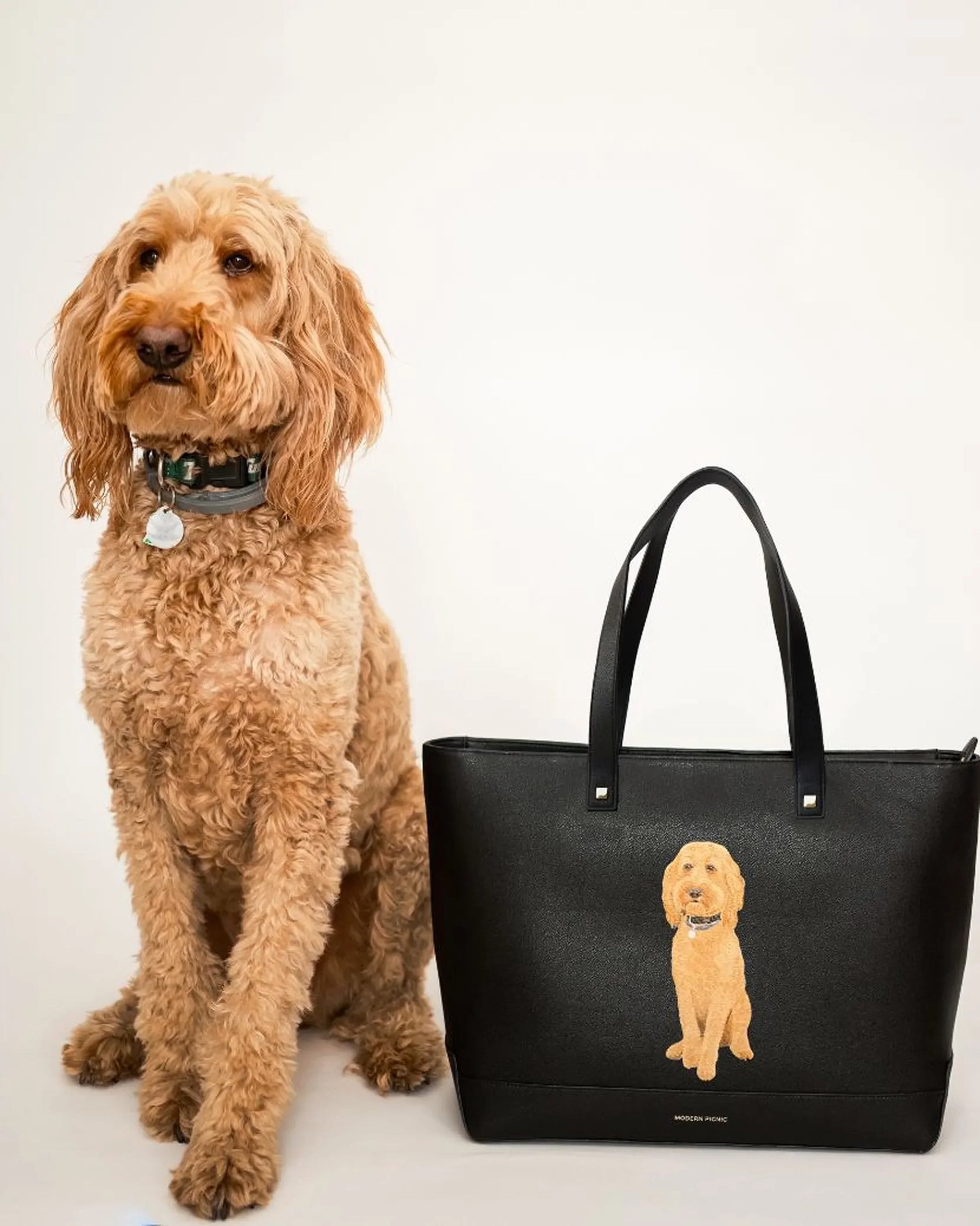 Curly-haired tan dog sitting beside a black vegan leather tote bag with matching dog illustration.