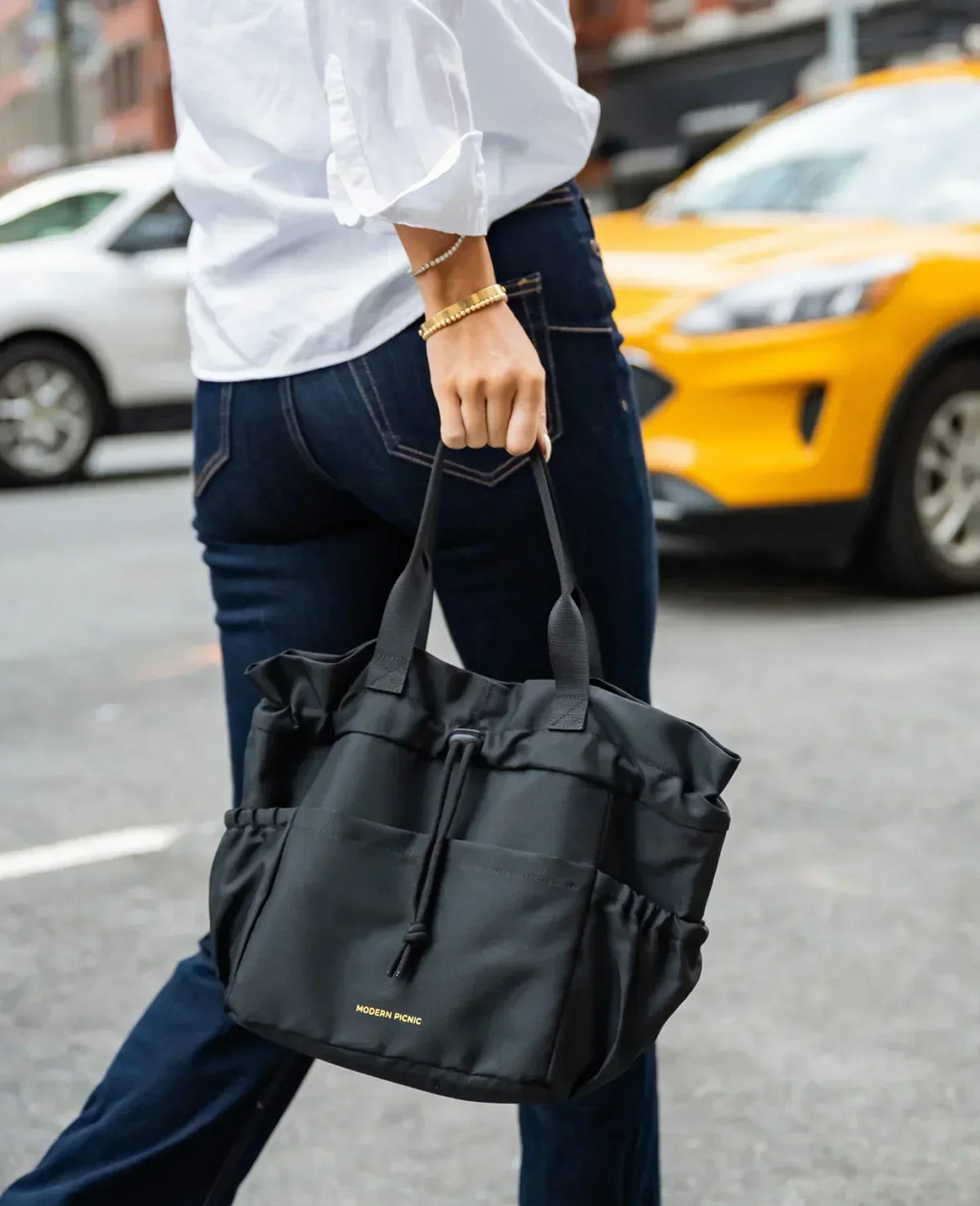 Woman in white shirt and blue jeans carrying a black Modern Picnic insulated tote bag.