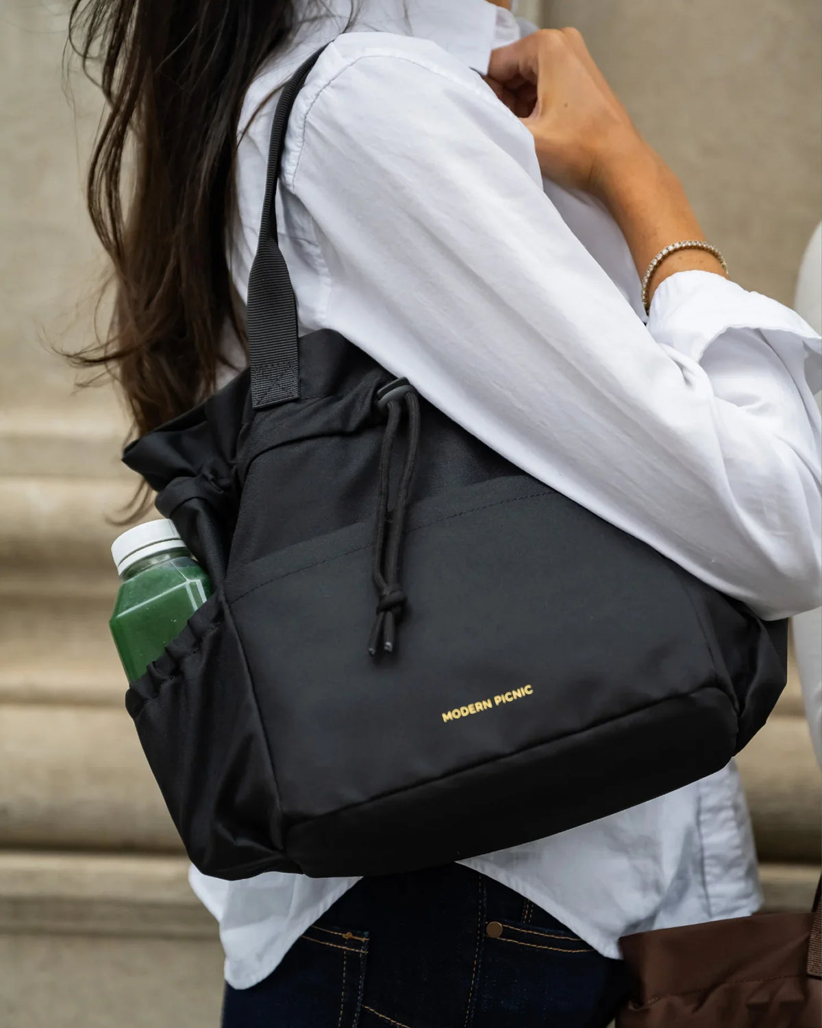 A woman wearing a white shirt carrying a black Modern Picnic insulated tote bag with a green bottle inside.