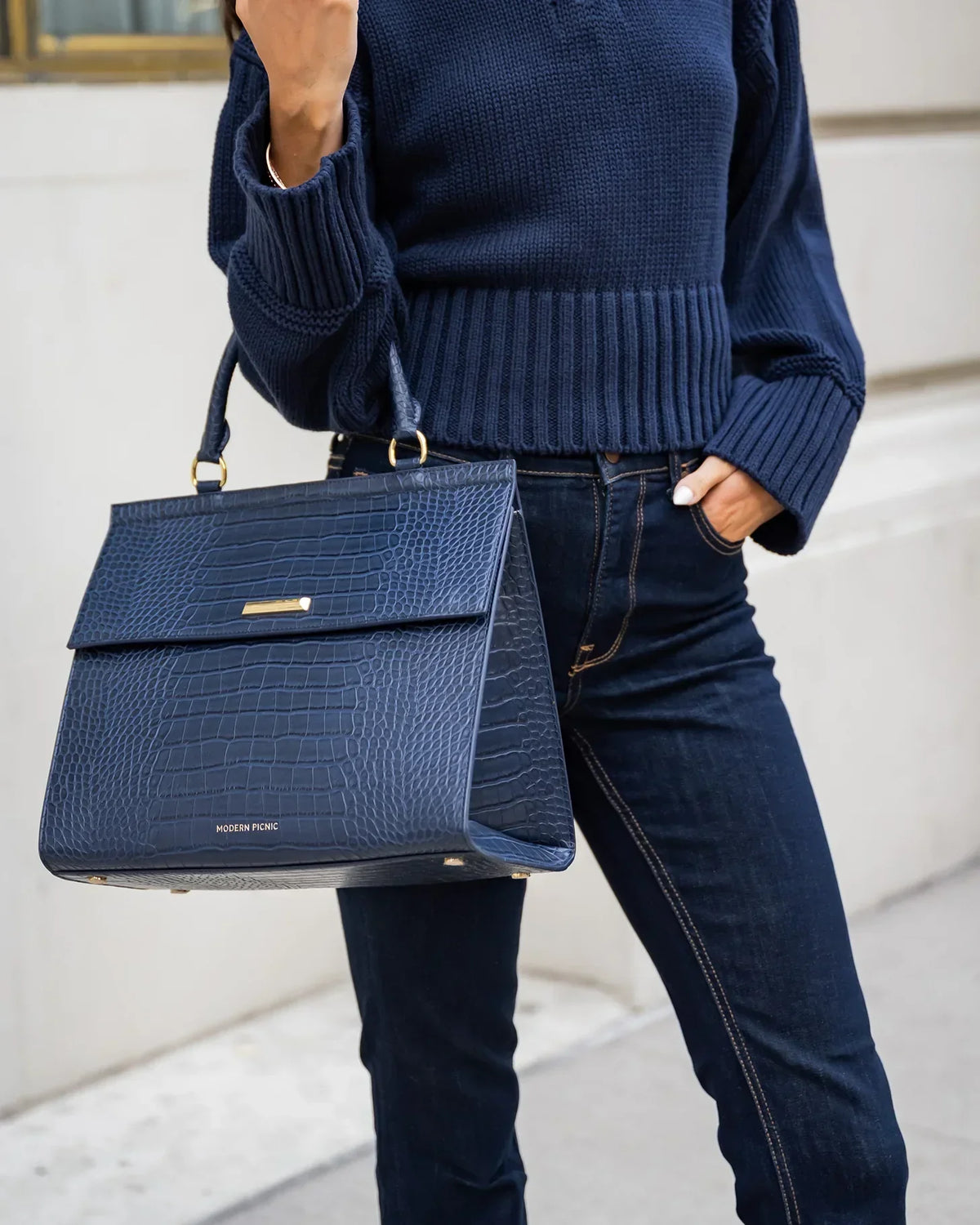 Woman wearing a navy sweater and jeans carrying a navy blue crocodile-embossed vegan leather tote.