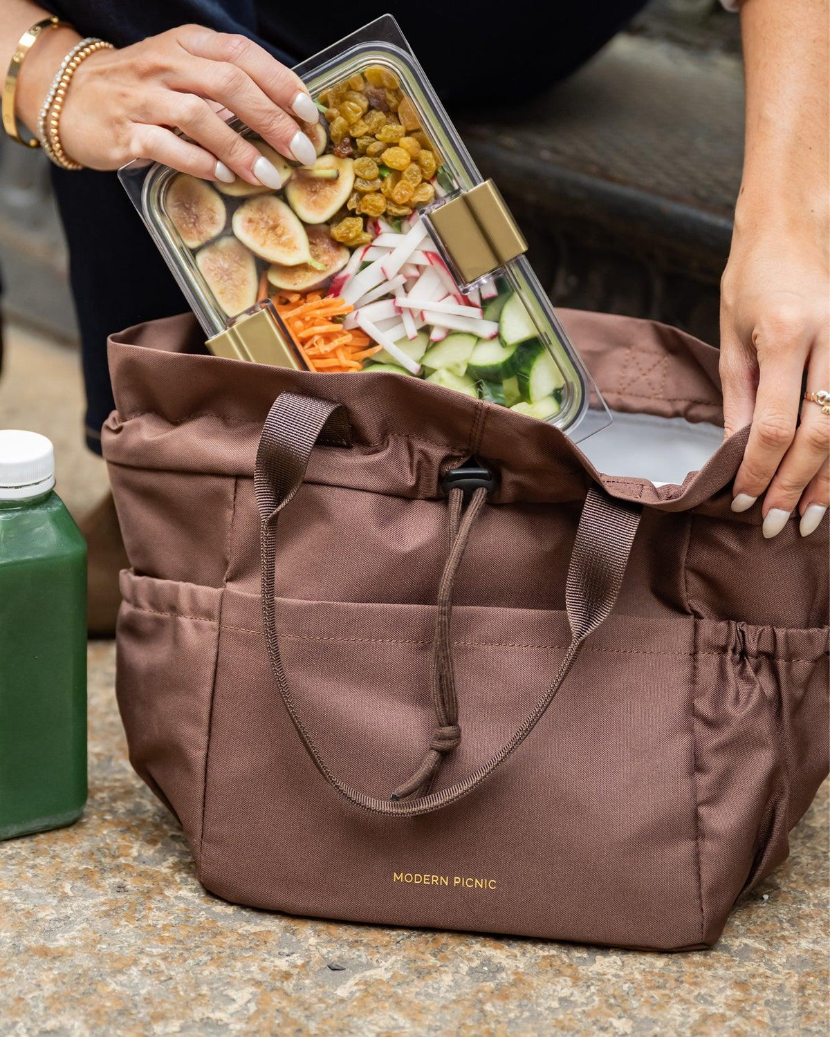 A person packing a clear glass meal prep container with fresh vegetables and fruit into a brown insulated lunch tote.