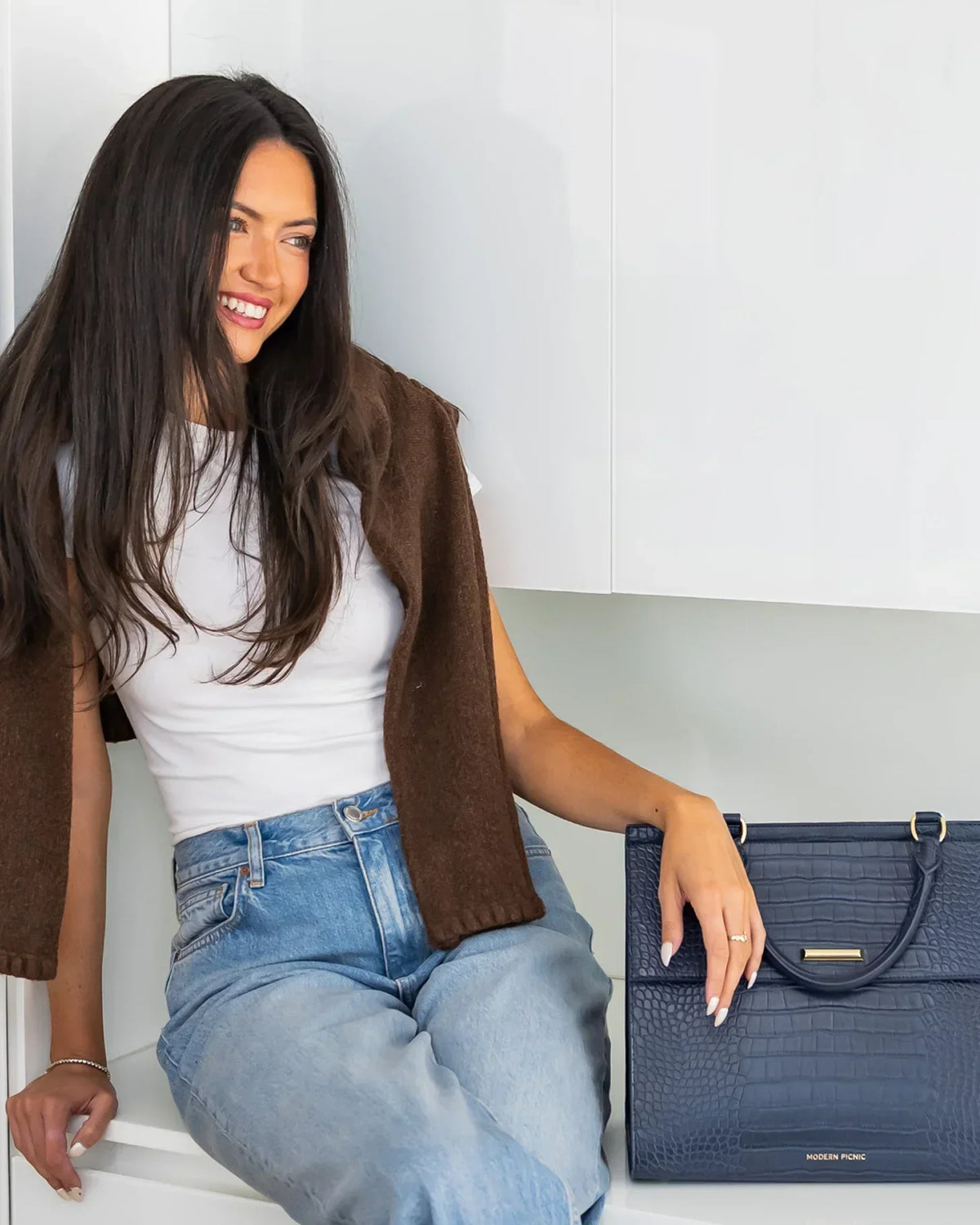 Woman in jeans and a white tee sitting next to a navy blue croc-embossed vegan leather tote bag.