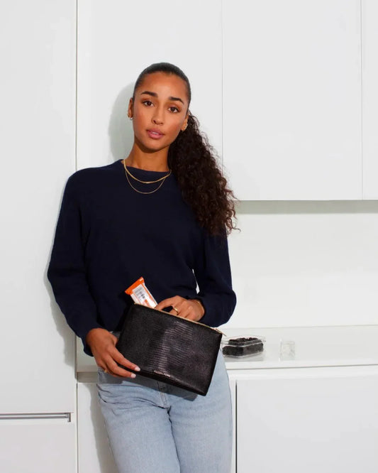 A woman with a long, curly ponytail, wearing a navy sweater and light-wash jeans, leans against white kitchen cabinets. She holds a small black pouch with a snack bar sticking out of the zipper.