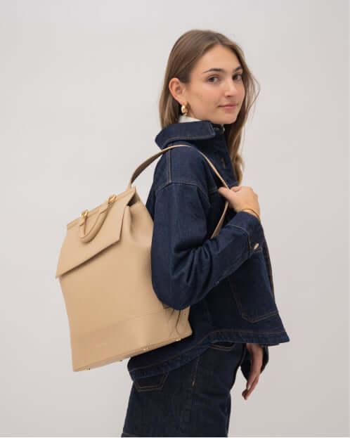 Woman wearing a denim jacket and gold jewelry, modeling the stylish Cream smooth vegan leather insulated lunch backpack over her shoulder.