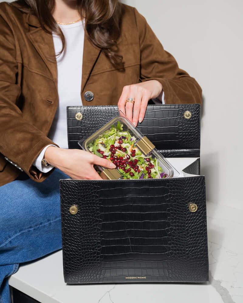 Woman placing salad container into black croc-texture insulated lunch bag, perfect for work or travel