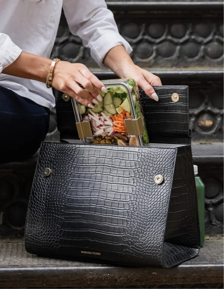 A person placing a healthy meal prep container into a black vegan leather insulated lunch bag.
